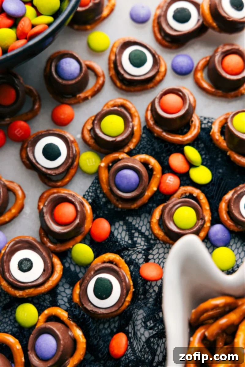Overhead view of two Monster Eyeball Halloween treats, made with pretzels, Rolos, M&M's, and large edible candy eyes, on a white surface.