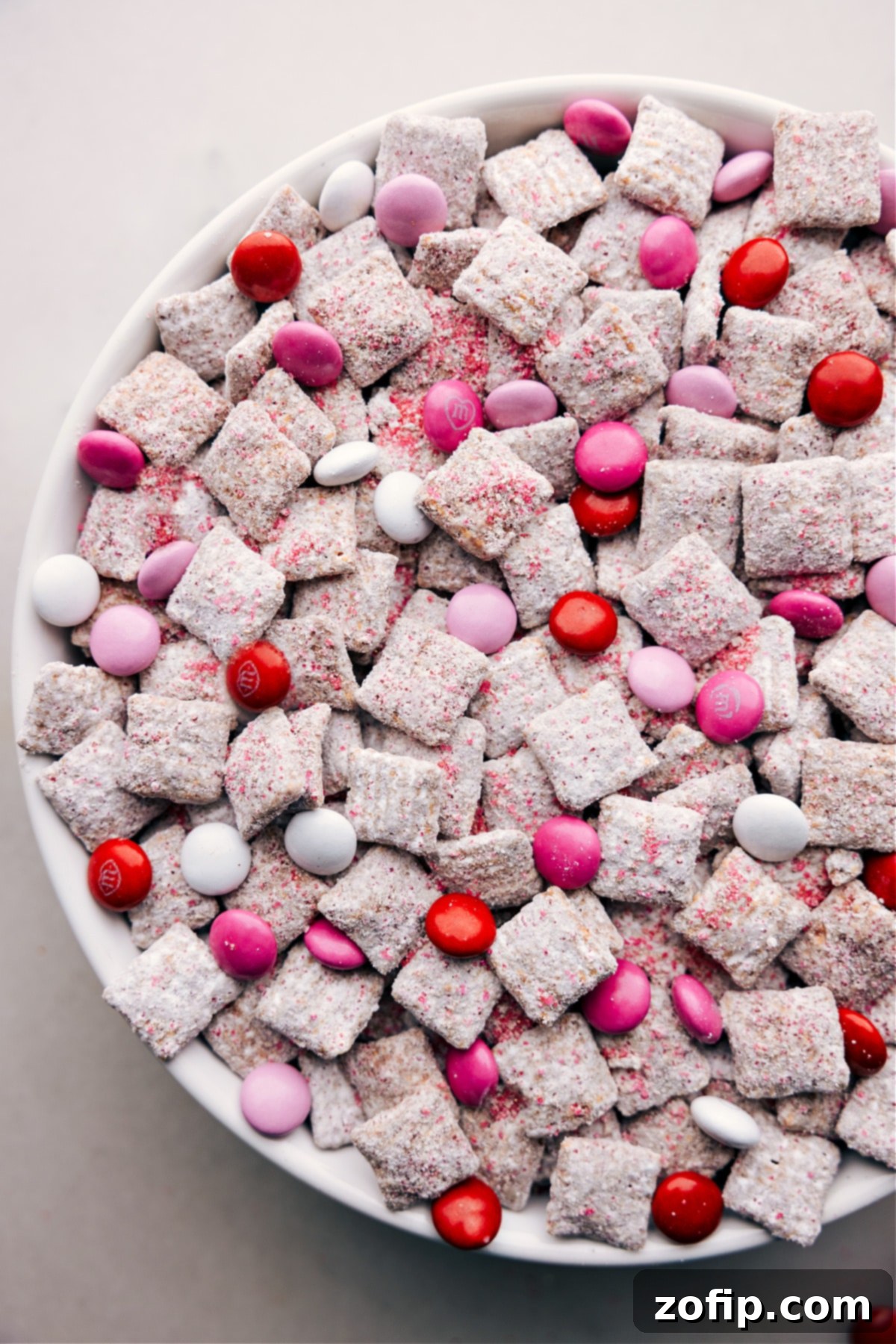 Our Favorite Valentine Muddy Buddies Recipe Close-up of Valentine Muddy Buddies in a bowl, showing the vibrant red, pink, and white M&M's interspersed throughout the powdered sugar-coated cereal.