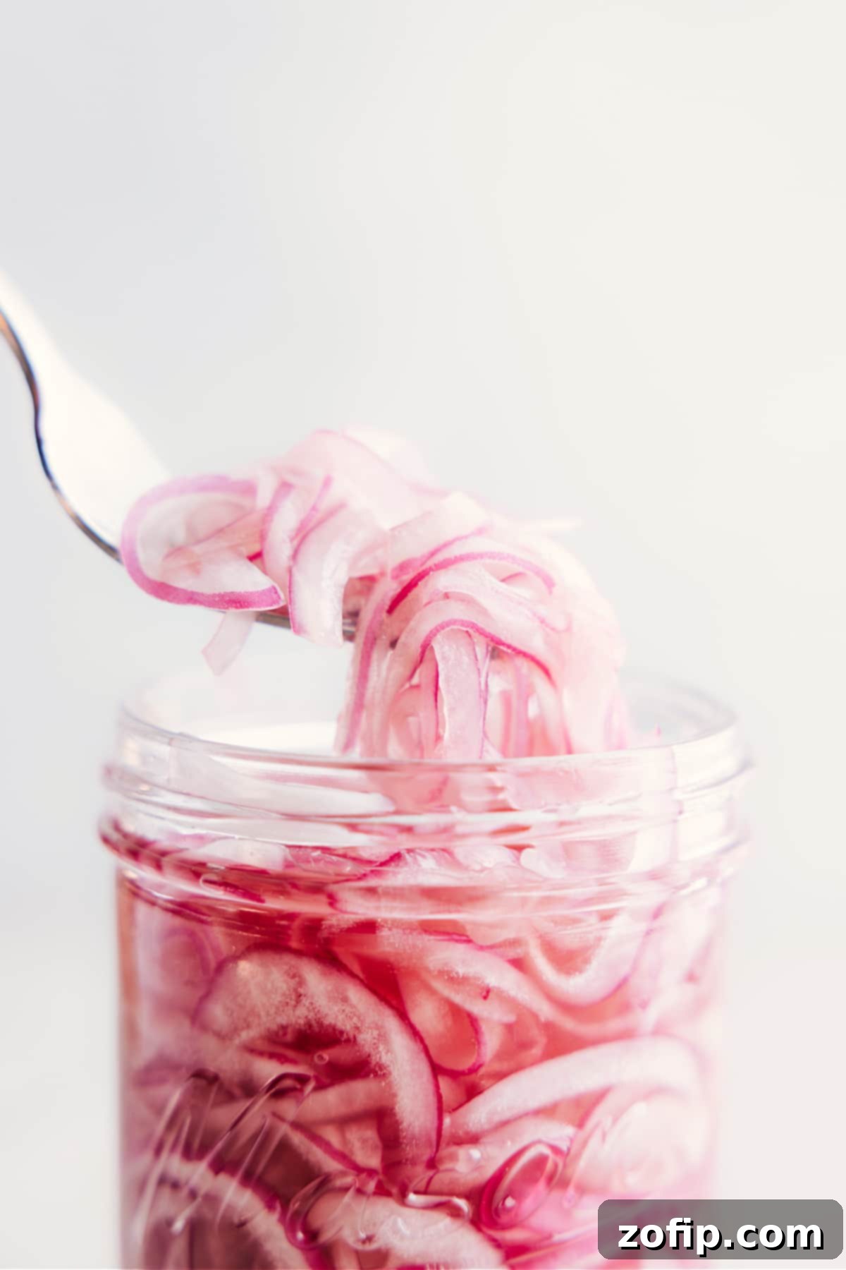 The Best Easy Pickled Red Onions A fork lifts a handful of perfectly pickled red onions from a glass jar, showcasing their vibrant color and texture.