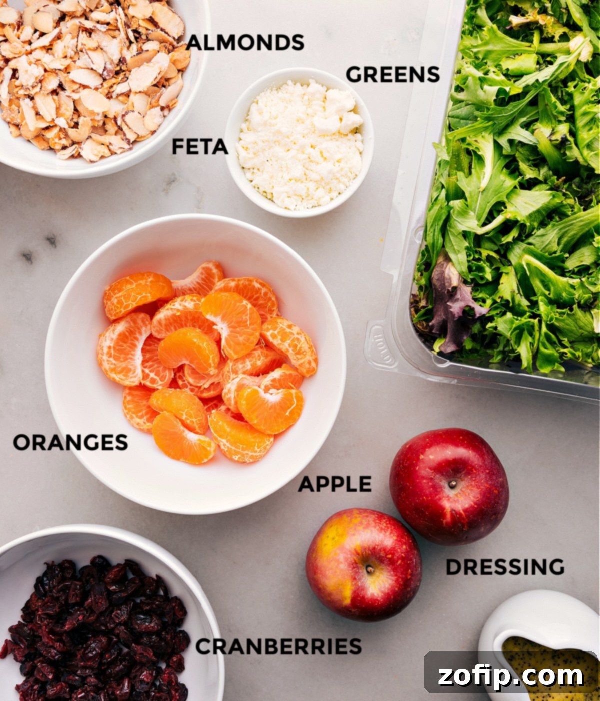 All fresh ingredients for Mandarin Orange Salad prepped on a wooden board, including a bowl of mixed greens, segmented clementines, sliced apples, dried cranberries, crumbled feta, and candied almonds.