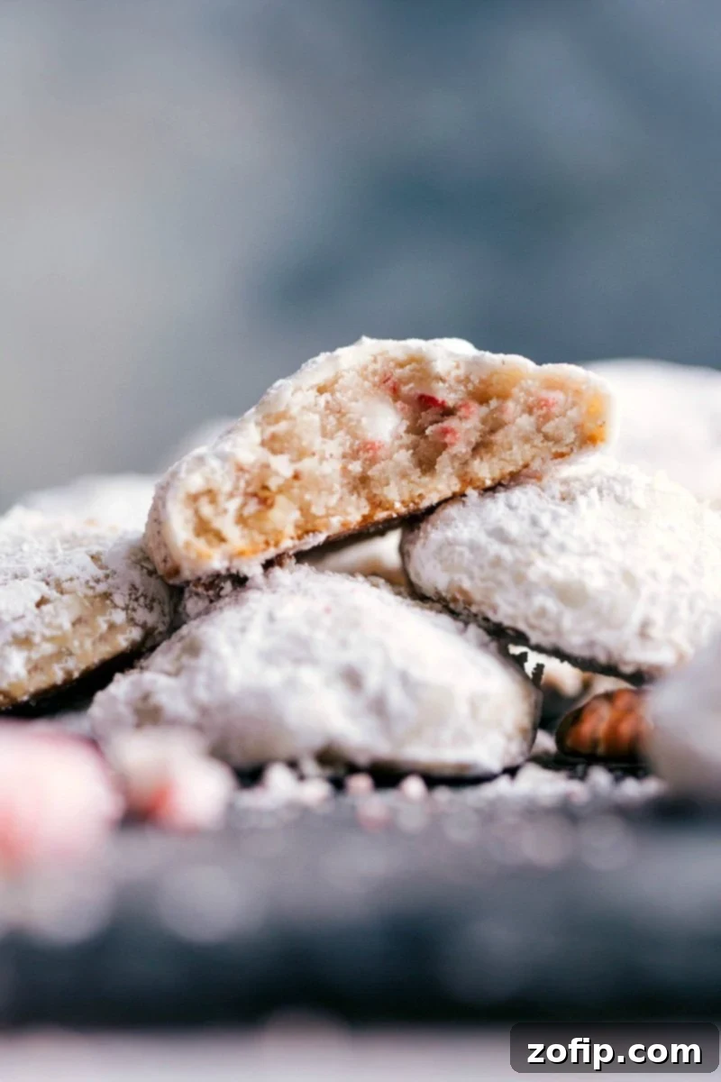 Delicious and seasonal peppermint snowball cookies stacked high, with one cookie showing a bite taken out, revealing its moist and flavor-packed interior with visible pecan and peppermint pieces.
