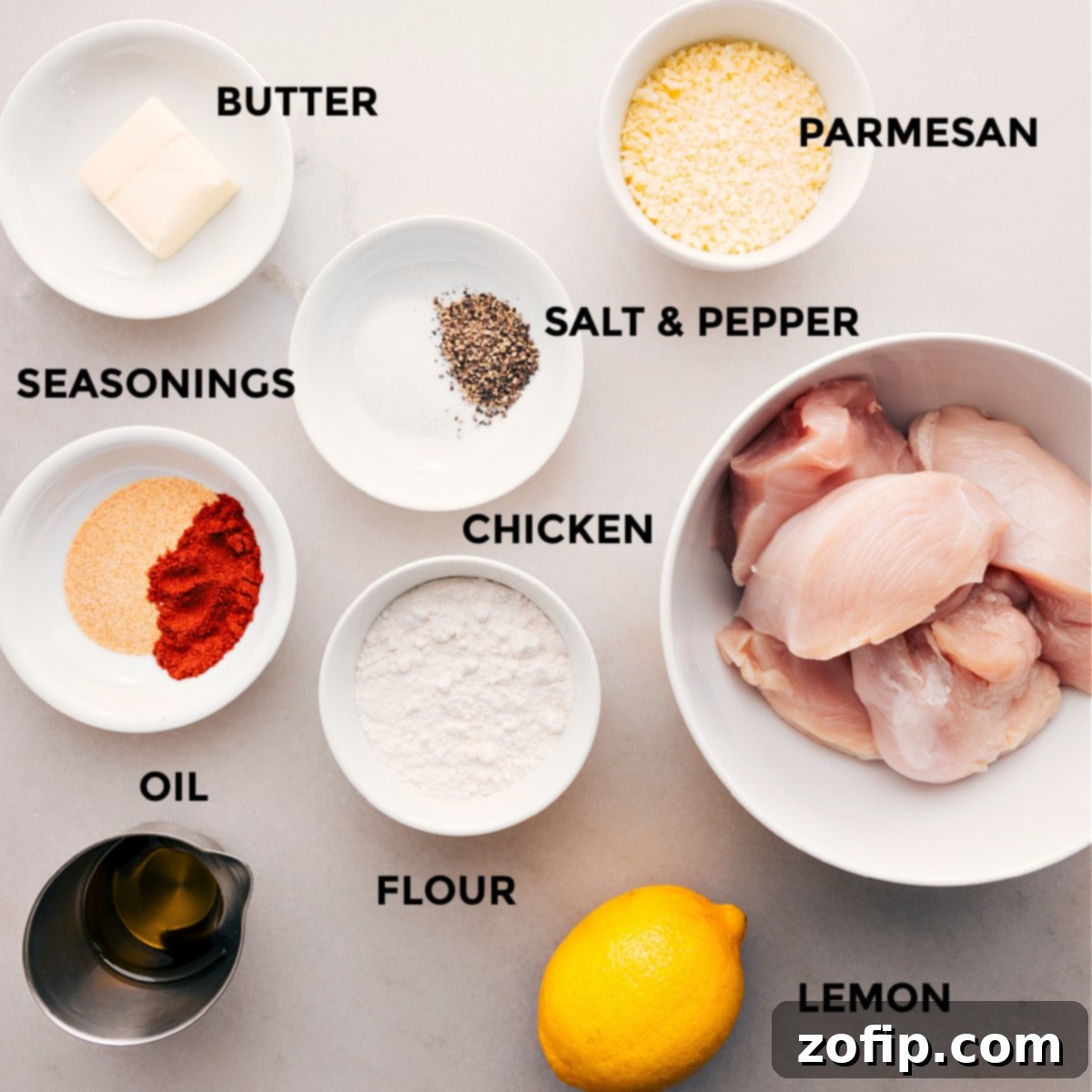 All the essential ingredients for making delicious homemade chicken cutlets are neatly arranged: fresh Parmesan cheese, a selection of savory seasonings, raw chicken breasts, all-purpose flour, olive oil, unsalted butter, and a bright lemon for serving.