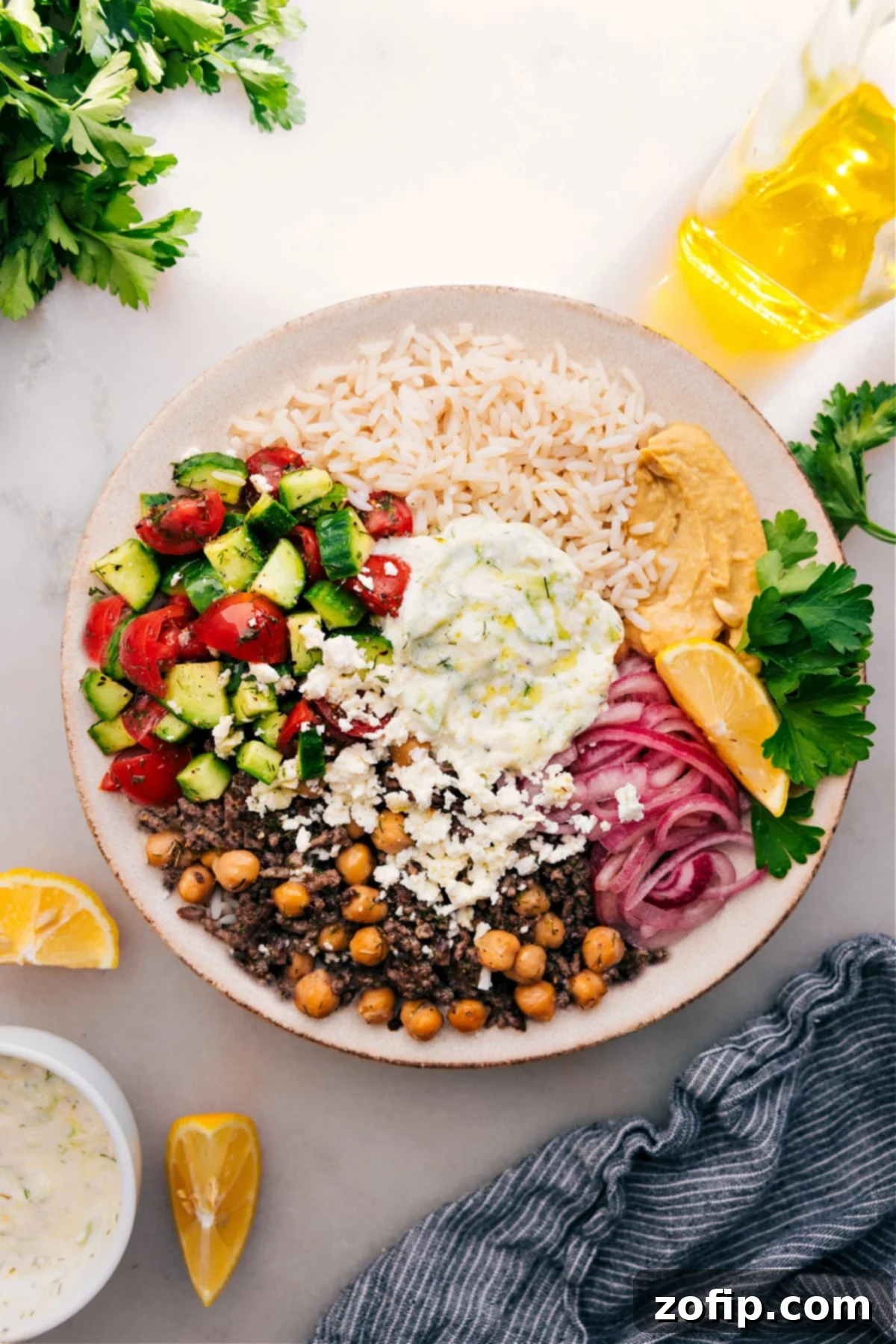 A colorful Greek Beef Bowl featuring seasoned ground beef, fluffy white rice, creamy tzatziki, fresh Greek salad, tangy pickled red onions, and smooth hummus.