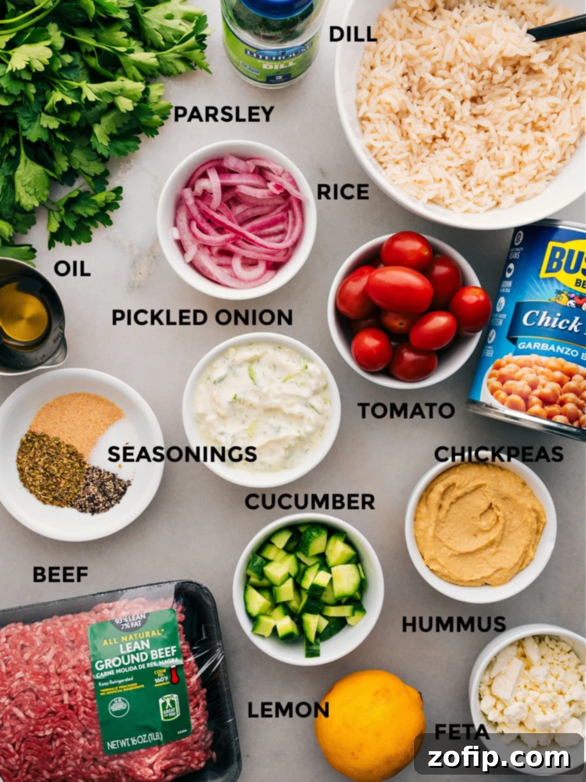 All the fresh ingredients for Greek Beef Bowls, prepped and ready for assembly: sliced red onions, cooked chickpeas, fluffy rice, chopped vegetables, a selection of seasonings, creamy hummus, fresh parsley, crumbled feta, and a lemon.