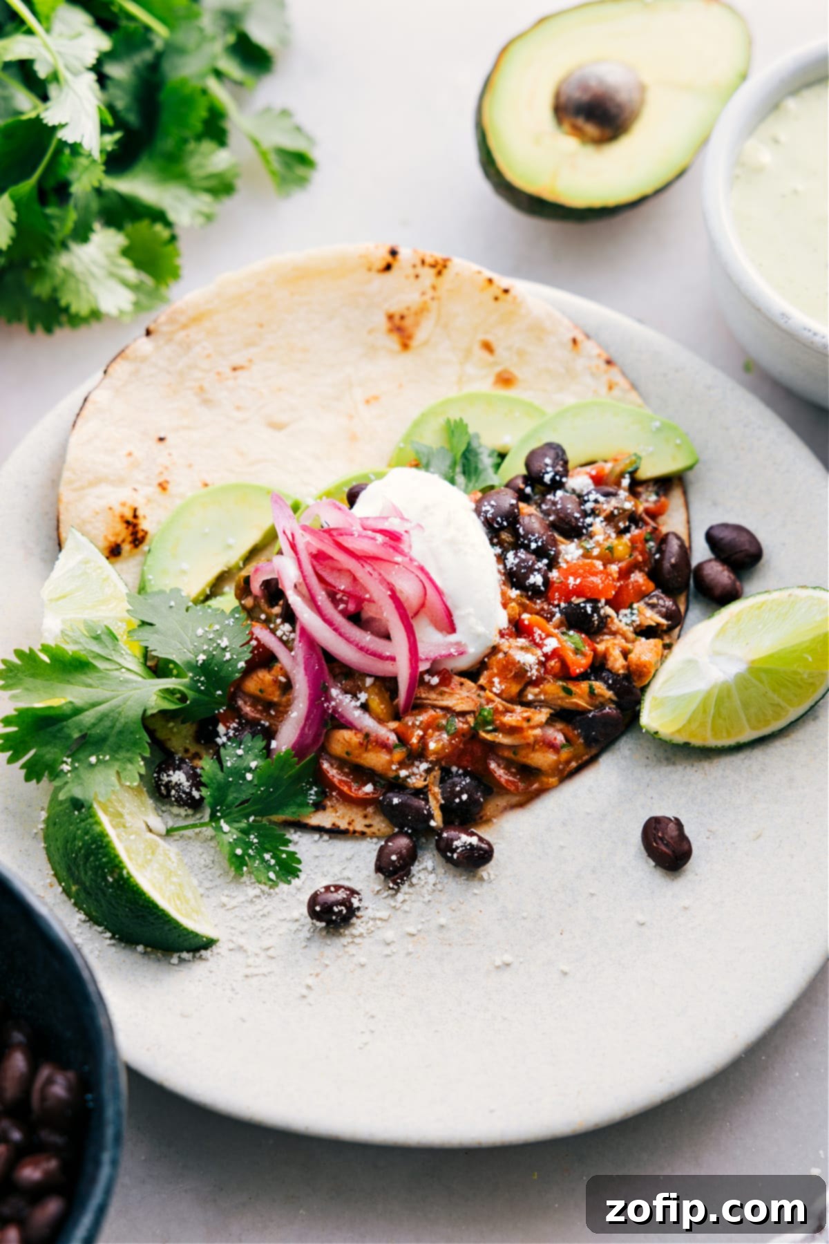 A perfectly assembled Crockpot Chicken Fajita in a warm tortilla, generously topped with black beans, sautéed onions, creamy sour cream, fresh avocado slices, shredded cheese, and a zesty lime wedge.