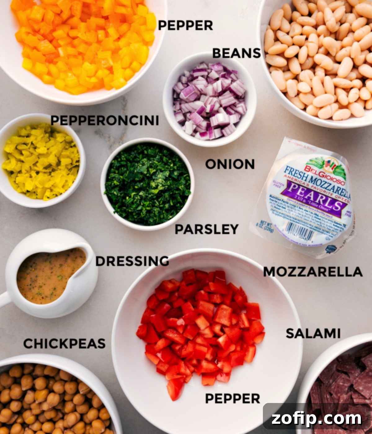 All the fresh ingredients for the Dense Bean Salad recipe are meticulously prepped and arranged, showcasing a colorful array of beans, diced onion, fresh parsley, mozzarella pearls, pepper salami, and sliced pepperoncini, ready for easy assembly.