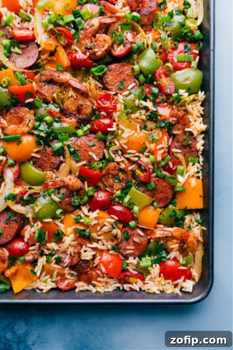 One-Pan Jambalaya: an easy, nutritious, 30-minute meal of oven-roasted & seasoned sausage, shrimp, veggies, and rice. Everything cooks together on one sheet pan! Recipe via ChelseasMessyApron #dinner #quick #easy #simple #best #veggies #shrimp #sausage #rice #onepan #jambalaya Overhead view of a vibrant One-Pan Jambalaya dish, showcasing sausage, shrimp, colorful bell peppers, onions, and rice, garnished with fresh green onions and parsley.