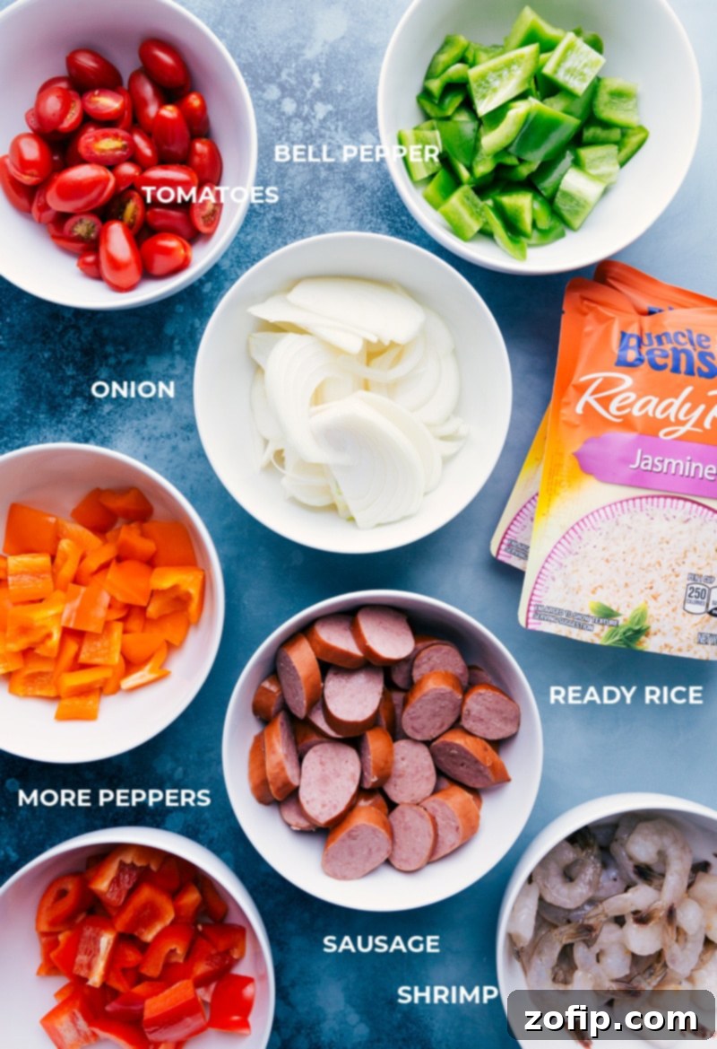 One-Pan Jambalaya features an easy, nutritious, 30-minute combination of oven-roasted & seasoned sausage, shrimp, veggies, and rice. Recipe via ChelseasMessyApron #dinner #quick #easy #simple #best #veggies #shrimp #sausage #rice #onepan #jambalaya A selection of fresh ingredients laid out, including colorful bell peppers, an onion, smoked sausage, raw shrimp, cherry tomatoes, and various spices, ready for the One-Pan Jambalaya.