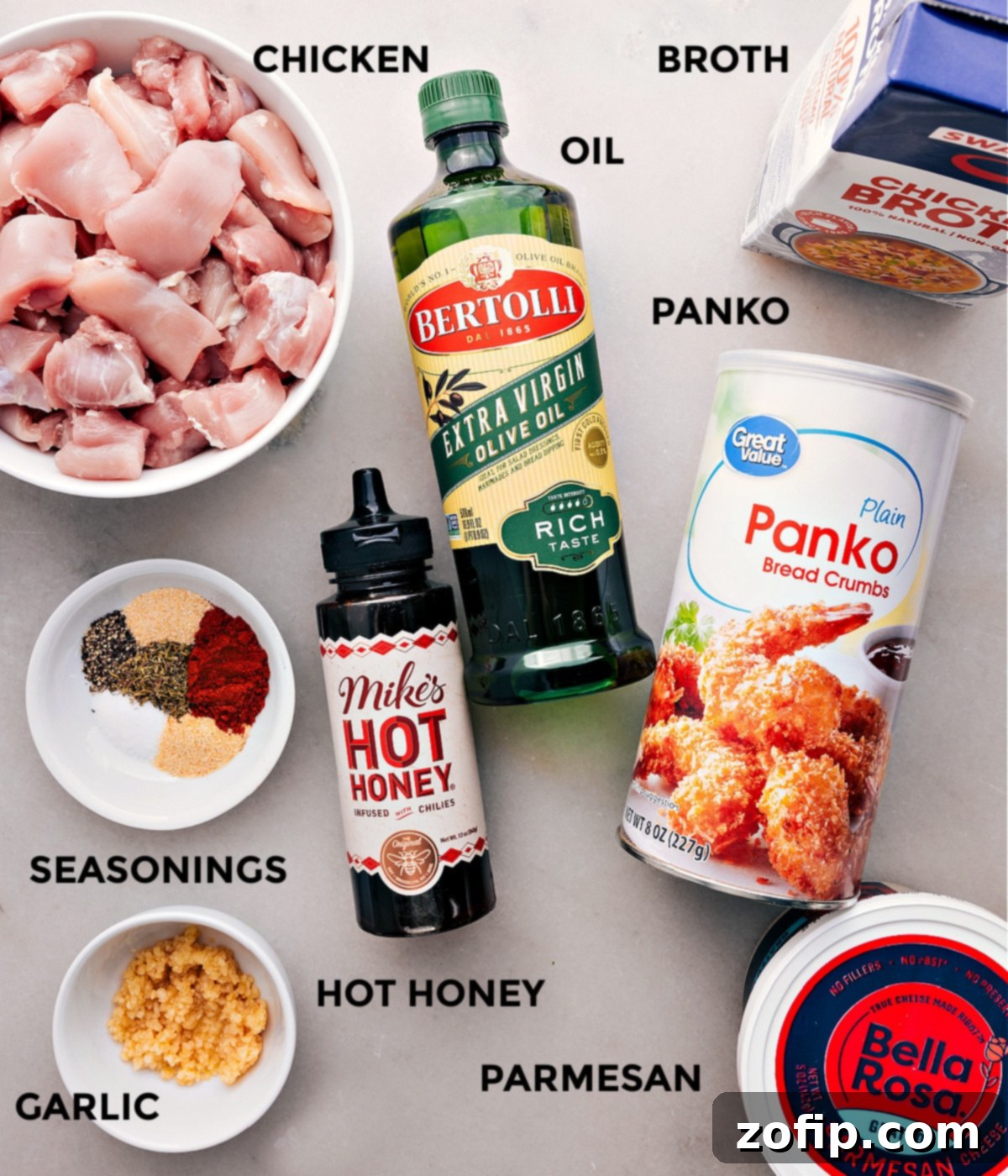 All the essential ingredients for hot honey chicken laid out: chicken thighs, olive oil, broth, minced garlic, spices, panko, Parmesan, butter, hot honey, and hot sauce.