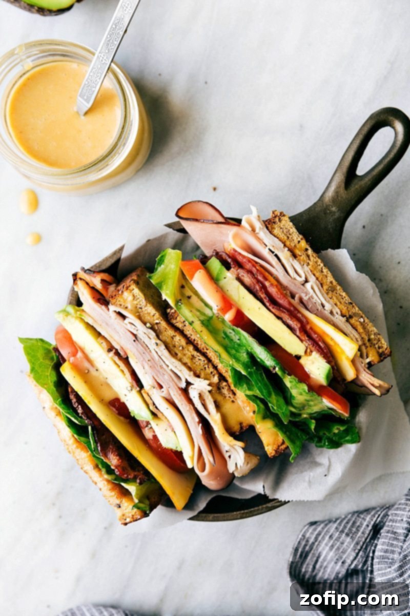 Overhead view of a prepared Honey Bacon Club Sandwich, showing the golden toasted bread, thick slices of bacon, deli meats, and vibrant vegetables, ready to be sliced and served.