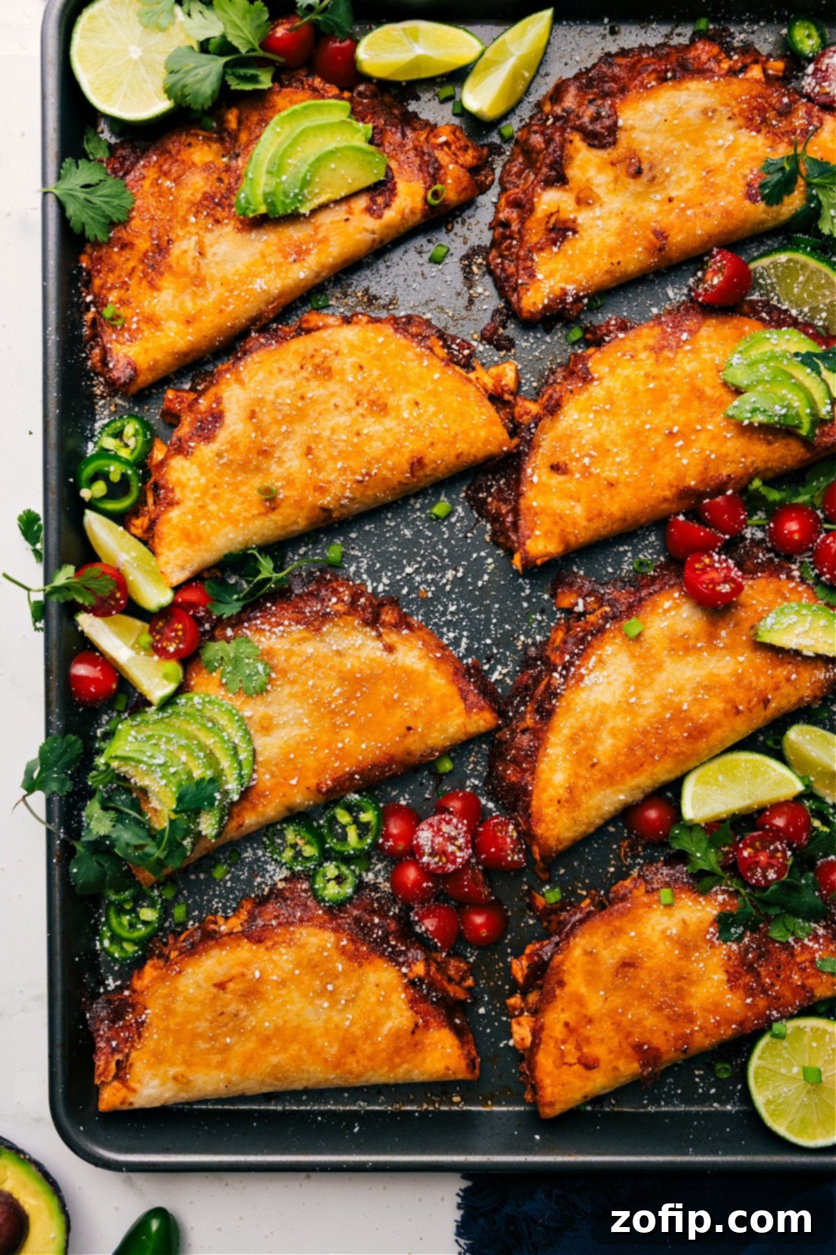 Golden-brown Crispy Baked Chicken Tacos arranged on a sheet pan, generously topped with fresh cilantro, vibrant diced tomatoes, and a drizzle of creamy chili-lime sauce, ready to be enjoyed.