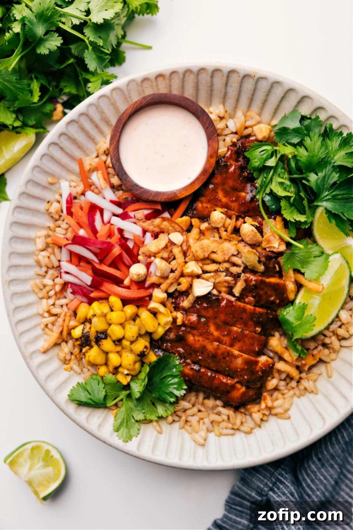 Asian Chicken Bowl served on brown rice, topped with corn, radish and carrot salad, limes, cilantro, and peanuts, with sauce on the side.