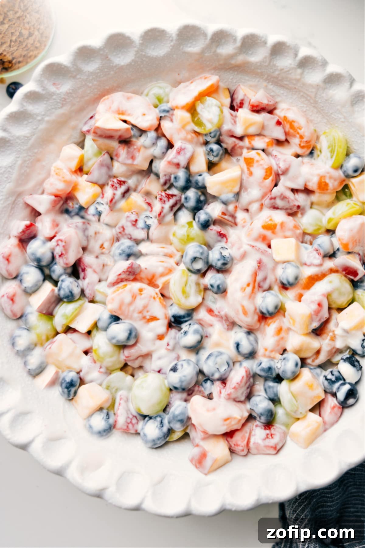 A vibrant fruit salad featuring an assortment of colorful fruits like strawberries, blueberries, grapes, mandarins, and apples, all gently coated in a rich, creamy white yogurt dressing and served in a large bowl, ready to be enjoyed.