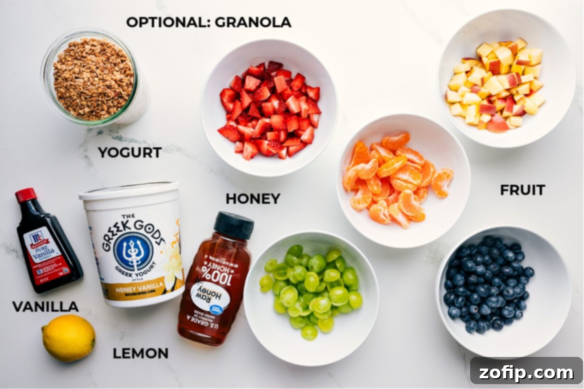 A flat lay photograph showcasing all the fresh and vibrant ingredients prepared for the fruit salad with yogurt, including mandarin segments, sliced strawberries, green grapes, blueberries, diced apples, a lemon, a bowl of honey vanilla Greek yogurt, and a small dish of honey and vanilla extract.
