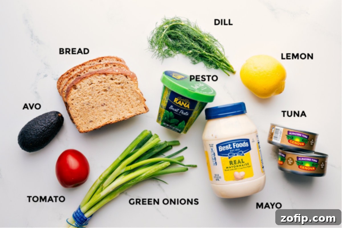 Ingredients In Tunacado All the fresh ingredients prepped for easy Tunacado assembly, including ripe avocado, sliced tomato, thin bread, chopped green onions, vibrant pesto, fresh dill, a lemon, high-quality tuna, and creamy mayonnaise.