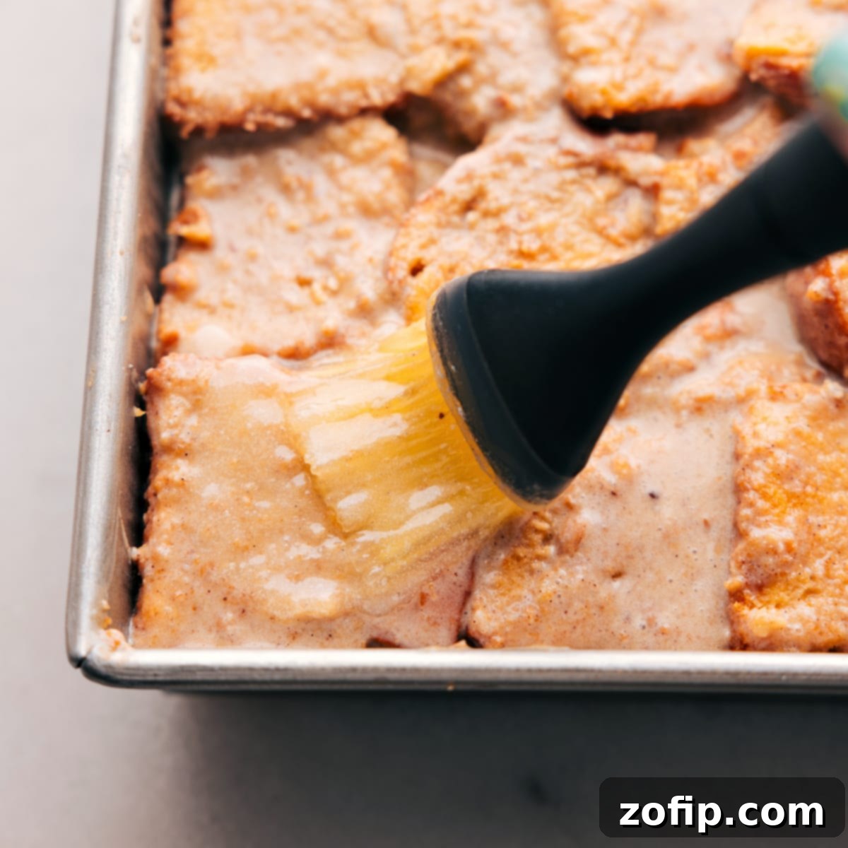 A baking pan filled with the raw bread pudding mixture, ready for the oven, with a pastry brush expertly applying melted butter across its top surface, ensuring a golden and crispy crust during baking.