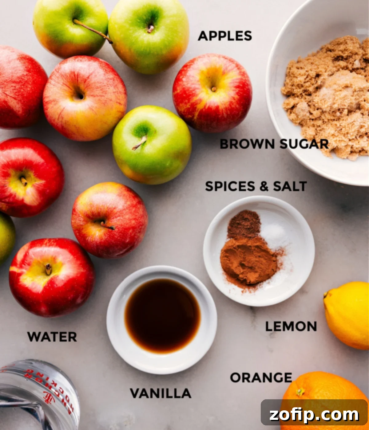 An array of fresh ingredients for hot apple cider including whole apples, an orange, brown sugar, cinnamon sticks, whole nutmeg, and a vanilla bean.