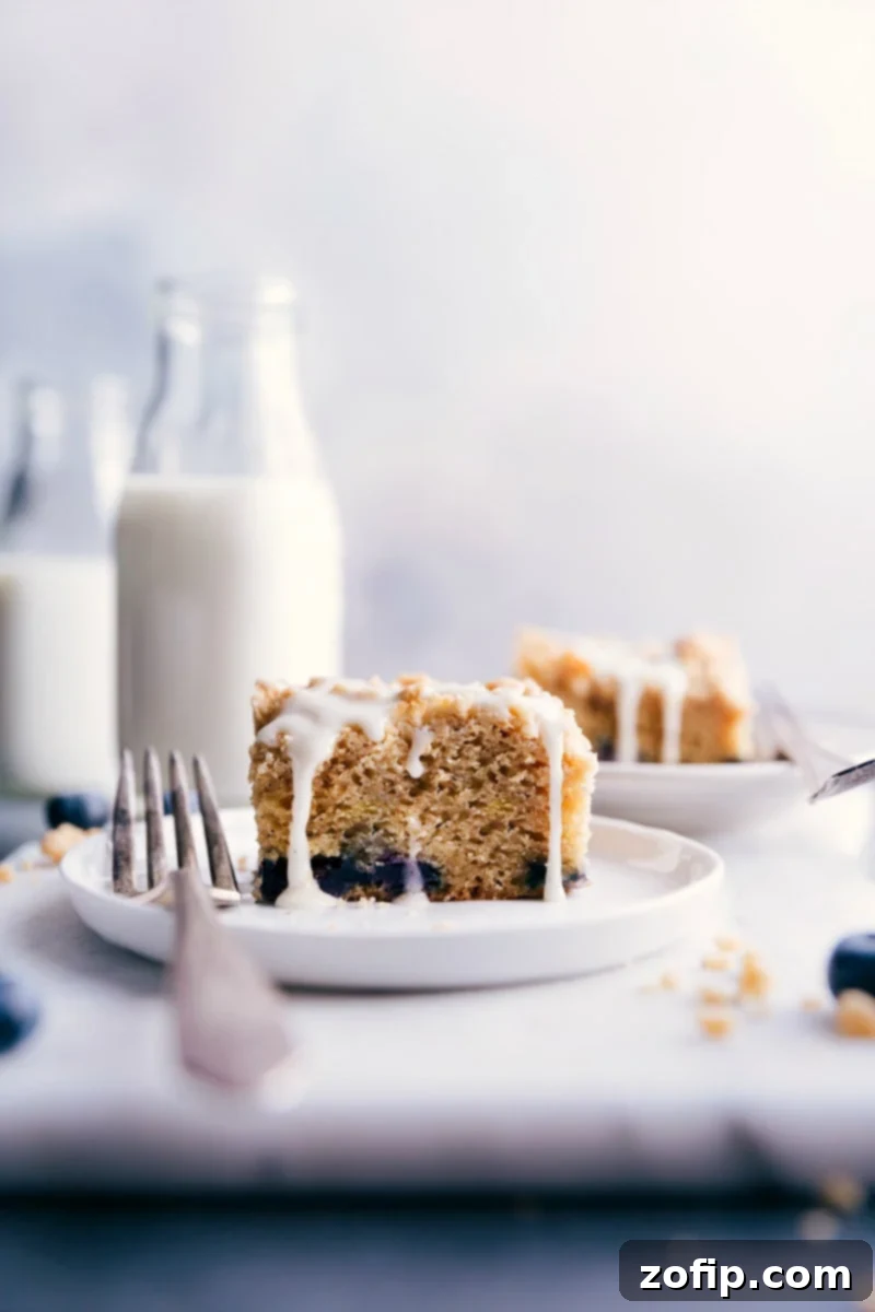 A slice of moist blueberry coffee cake on a white plate, showcasing the streusel topping and lemon glaze, with a fork beside it.