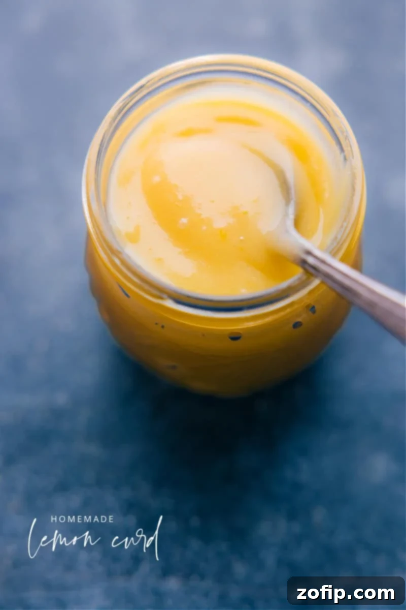 Homemade Lemon Curd: A rich, tangy, and versatile citrus spread for all your baking and breakfast needs. Overhead image of bright yellow Lemon Curd in a clear glass jar, with a silver spoon resting inside, invitingly thick and glossy.