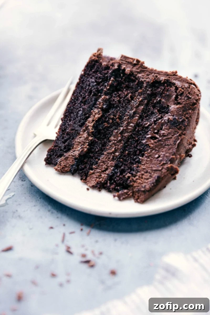 A perfectly sliced piece of moist and rich chocolate cake, layered with creamy triple chocolate buttercream, sits on a white plate, ready for an exquisite indulgence. The texture is inviting, hinting at its ultimate deliciousness.
