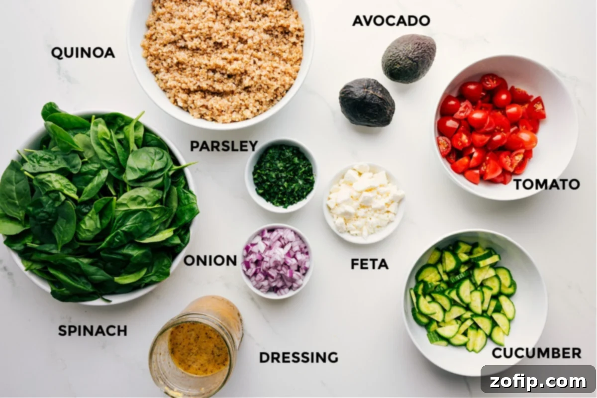 All the fresh ingredients for Quinoa Salad prepped and laid out on a cutting board, including cooked quinoa, chopped avocado, cherry tomatoes, feta cheese, cucumber, fresh parsley, red onion, a jar of lemon dressing, and baby spinach.