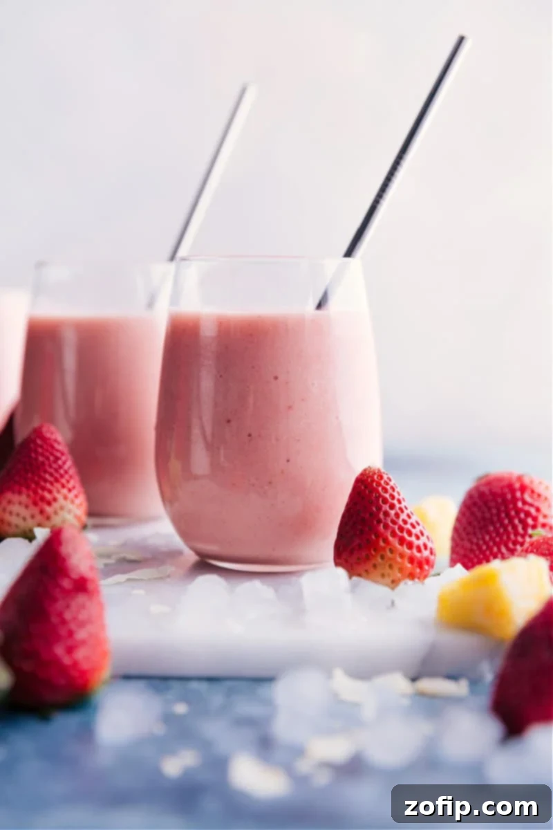 Homemade Strawberry Piña Colada Smoothie: A creamy, sweet, and perfectly blended non-alcoholic drink featuring coconut, pineapple, and strawberries. Two vibrant Strawberry Piña Colada smoothies in tall glasses, garnished with fresh strawberries and pineapple wedges, sitting on a light-colored surface, evoking a tropical and refreshing vibe.