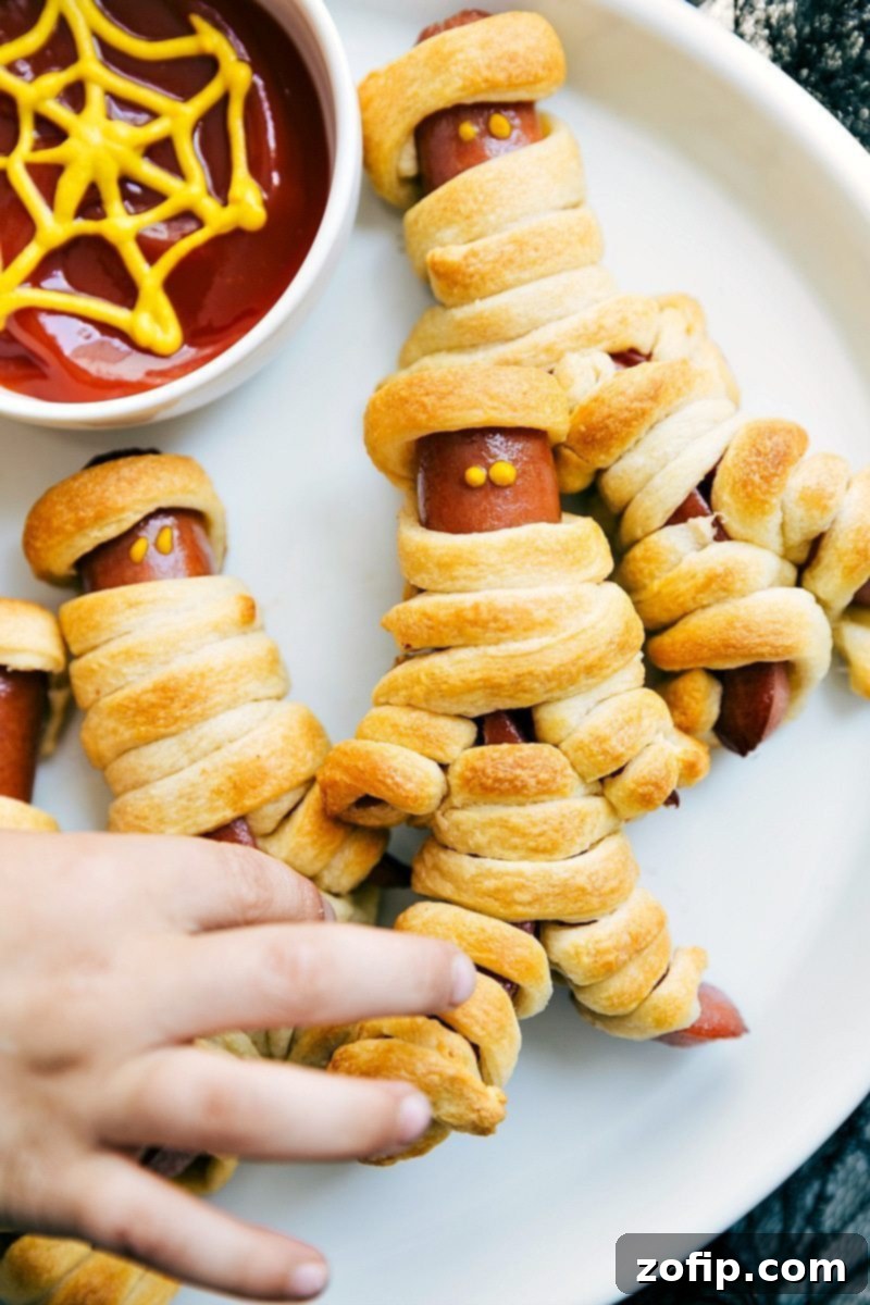 Four easy, & creative Halloween party appetizers that are so delicious -- Spiderweb 7-Layer Dip, Walking Dead Mummy Dogs, Monster Meatball Sliders, & Cheesy Monster Fingernail Bread. chelseasmessyapron.com #mummy #hotdog #mustard #halloween #treats #appetizer Image of spooky Walking Dead Mummy Dogs, ideal for kid-friendly Halloween appetizers
