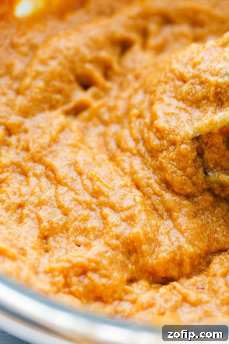 Canned pumpkin puree being added to the creamy batter mixture in a mixing bowl, enriching the cupcake base with autumnal flavor.