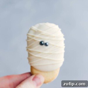 Ghoulish Goodies 2 An adorable and festive mummy cookie being held, decorated with white icing and eyes to resemble a mummy, perfect for Halloween.