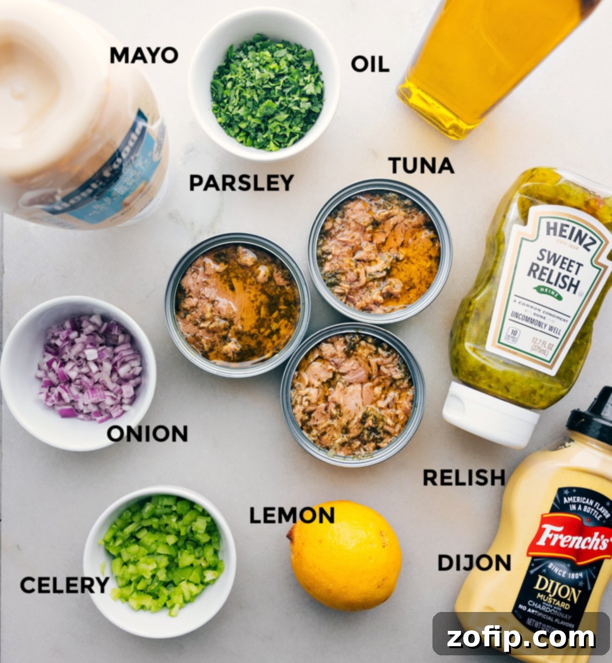 All the fresh ingredients laid out on a clean surface, including jars of mayo, a bowl of parsley, olive oil, cans of tuna, a chopped onion, pickle relish, a lemon, Dijon mustard, and diced celery, ready for assembly.