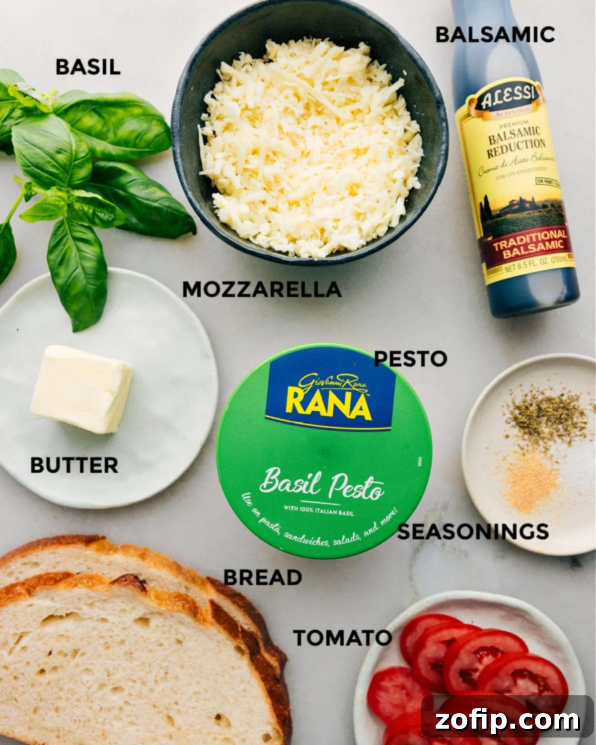 The vibrant and fresh ingredients for Caprese Grilled Cheese laid out: basil leaves, balsamic reduction, shredded mozzarella, basil pesto, softened butter, garlic powder, Italian seasoning, sliced Roma tomatoes, and thick-cut sourdough bread.