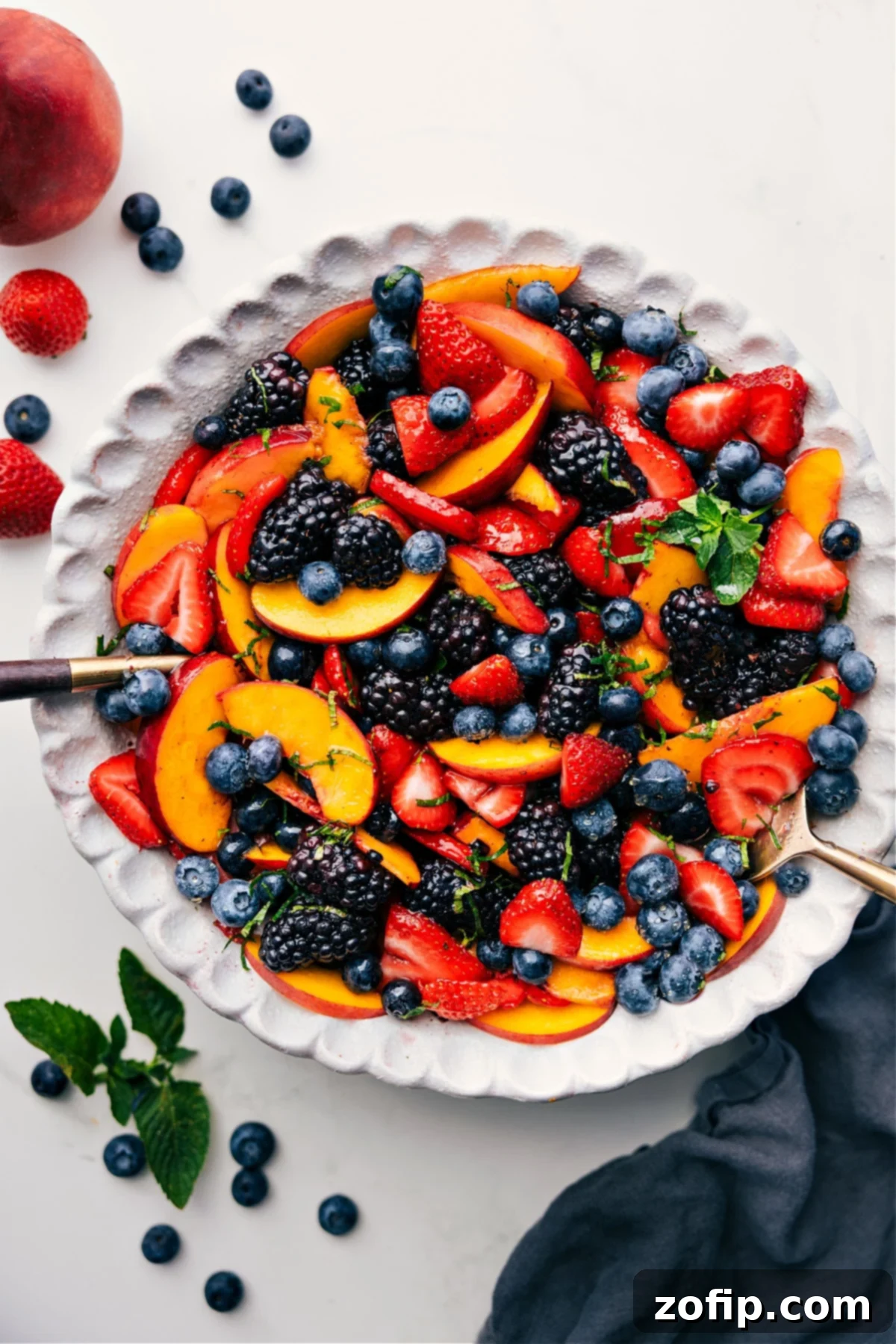A beautifully arranged Peach Fruit Salad featuring sliced peaches, blueberries, blackberries, and strawberries, glistening with a light dressing and garnished with fresh mint.