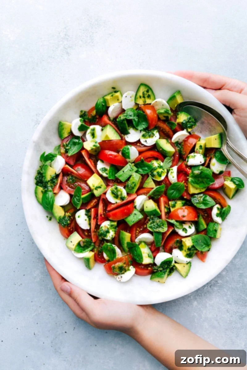 A vibrant Caprese Salad in a bowl, generously tossed with basil vinaigrette and garnished with fresh torn basil leaves and creamy avocado slices.