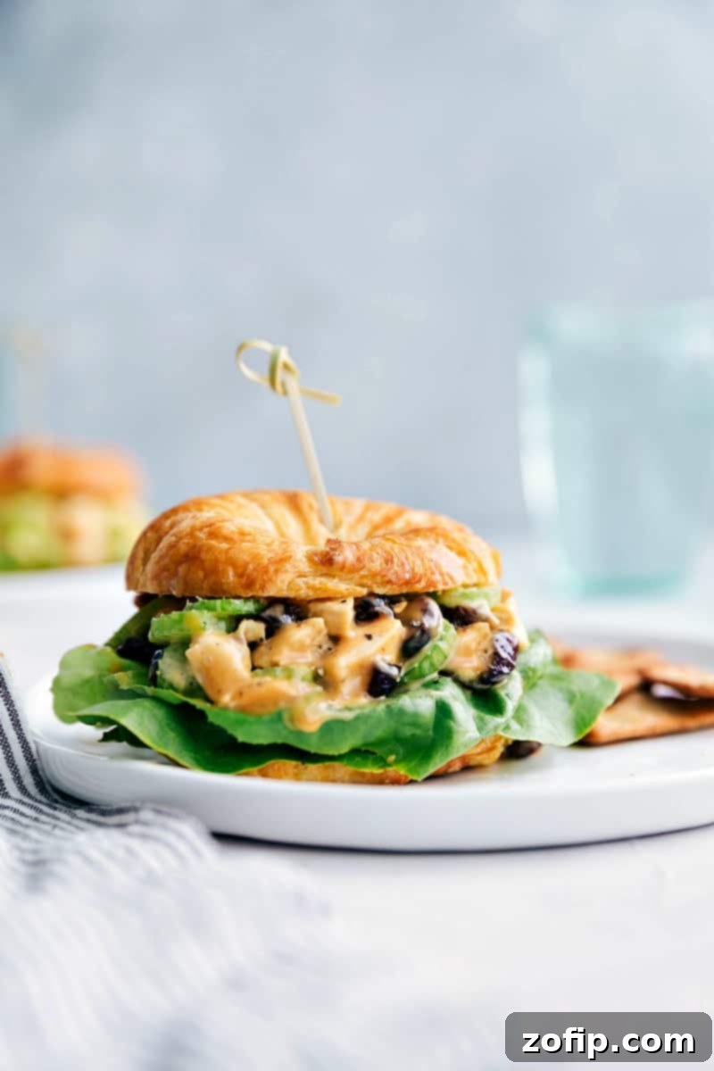 Close-up shot of a Honey Mustard Chicken Salad Sandwich on a croissant with crisp crackers and a glass of water in the background.