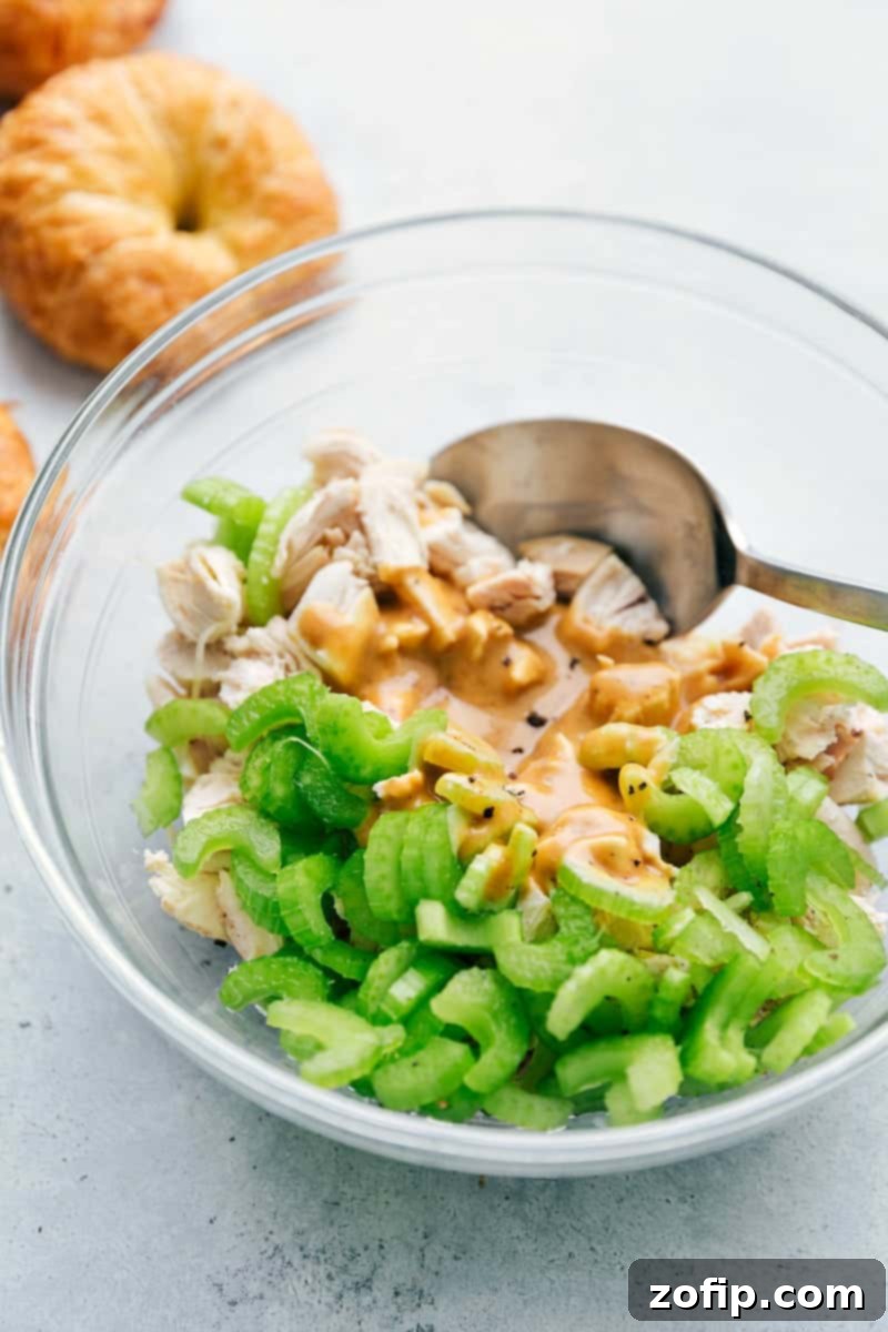 A bowl of Honey Mustard Chicken Salad mixture being stirred, with fresh croissant rolls awaiting in the background.