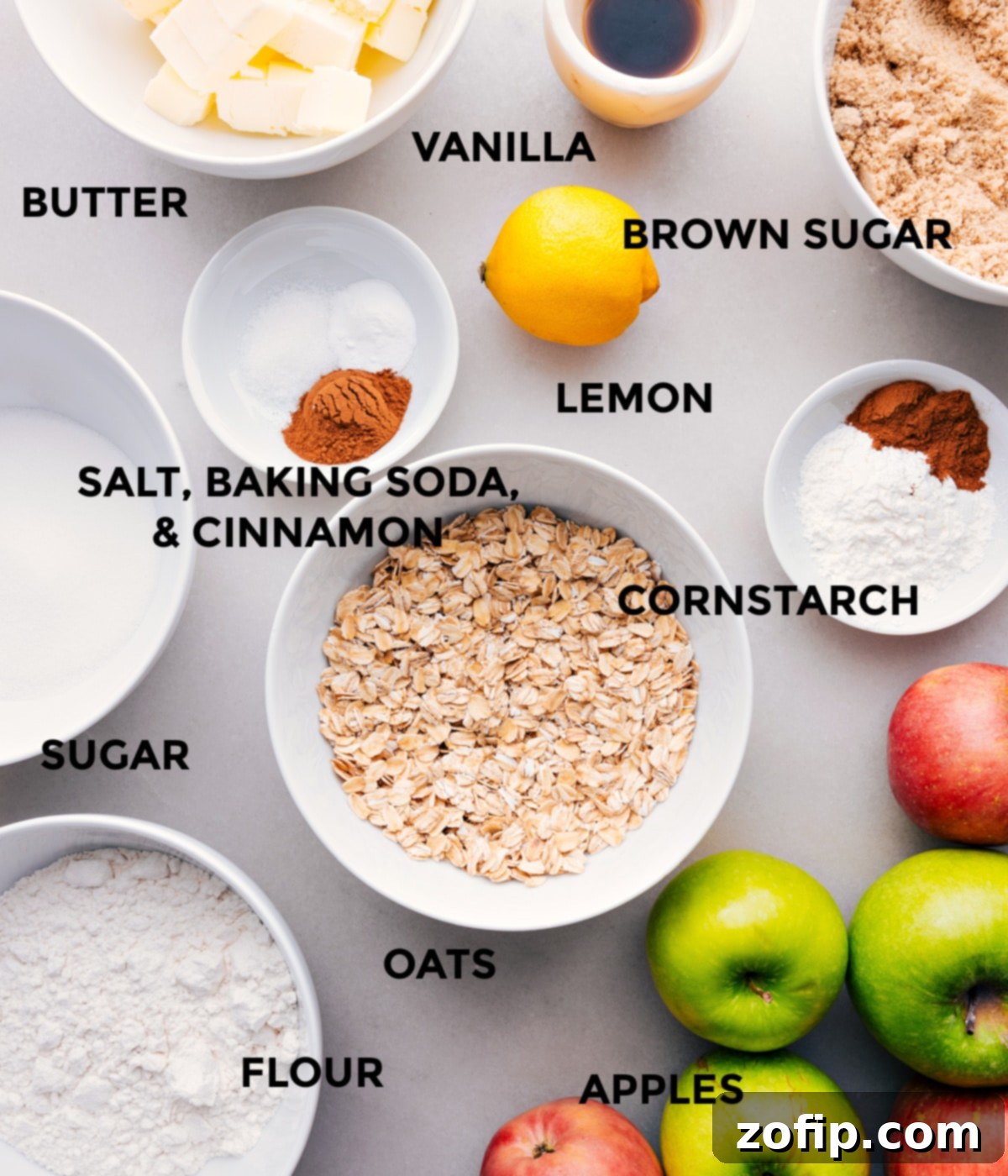 Assorted ingredients for apple crumble laid out on a wooden surface: fresh apples, butter, brown sugar, flour, oats, cinnamon, and lemon.