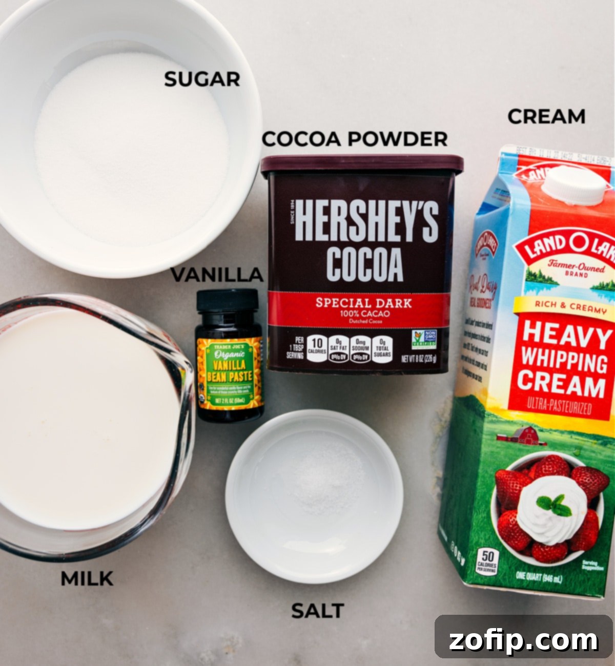 A selection of high-quality ingredients laid out on a kitchen counter, ready for preparing homemade hot chocolate, featuring Dutch-process cocoa, sugar, salt, milk, heavy cream, and vanilla extract.