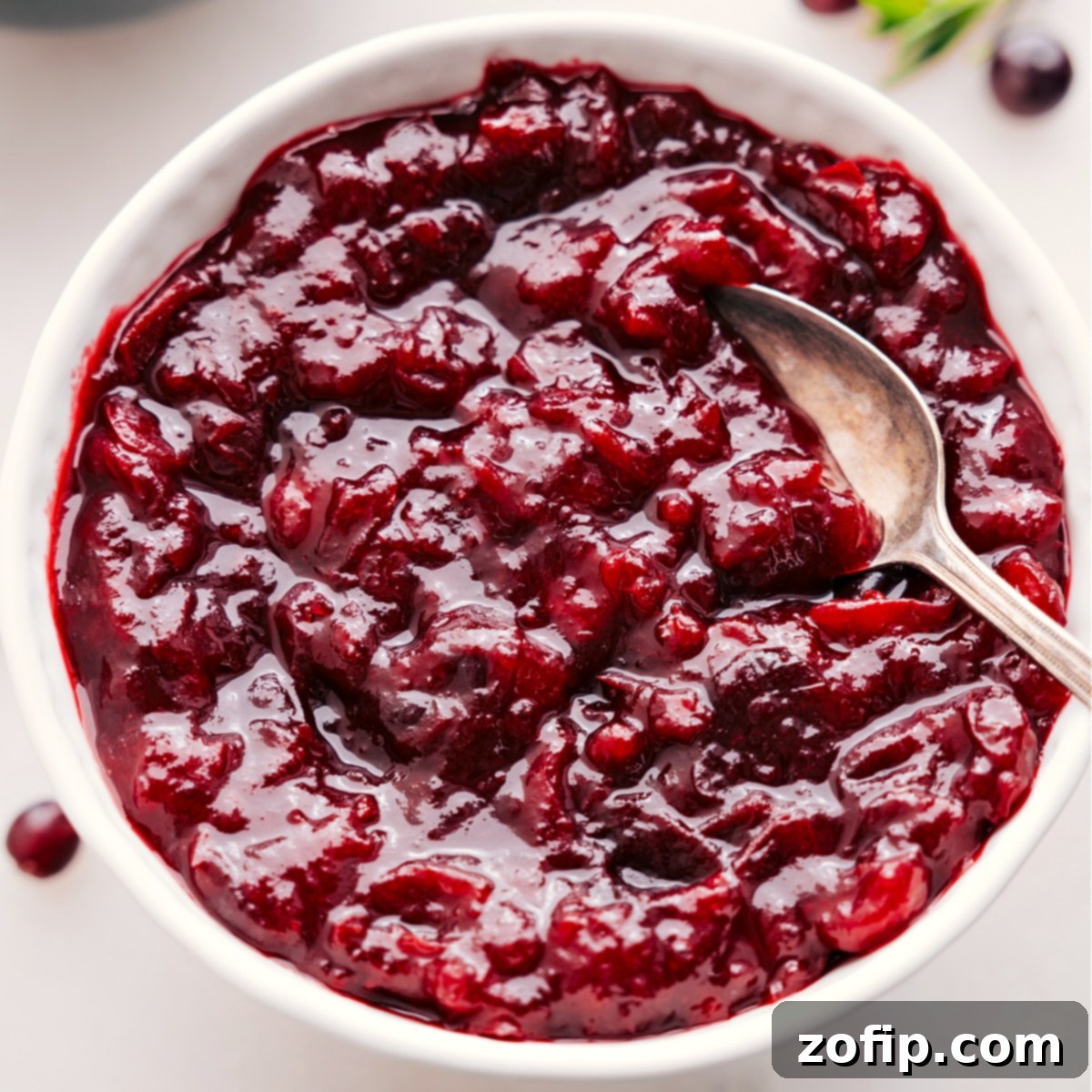 A bowl of glossy homemade cranberry sauce, ready to be served as part of a lavish holiday meal, with a spoon nestled in it.