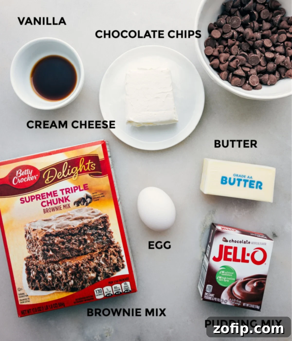 All the essential brownie cookie ingredients laid out, including brownie mix, butter, cream cheese, egg, vanilla, pudding mix, and chocolate chips.