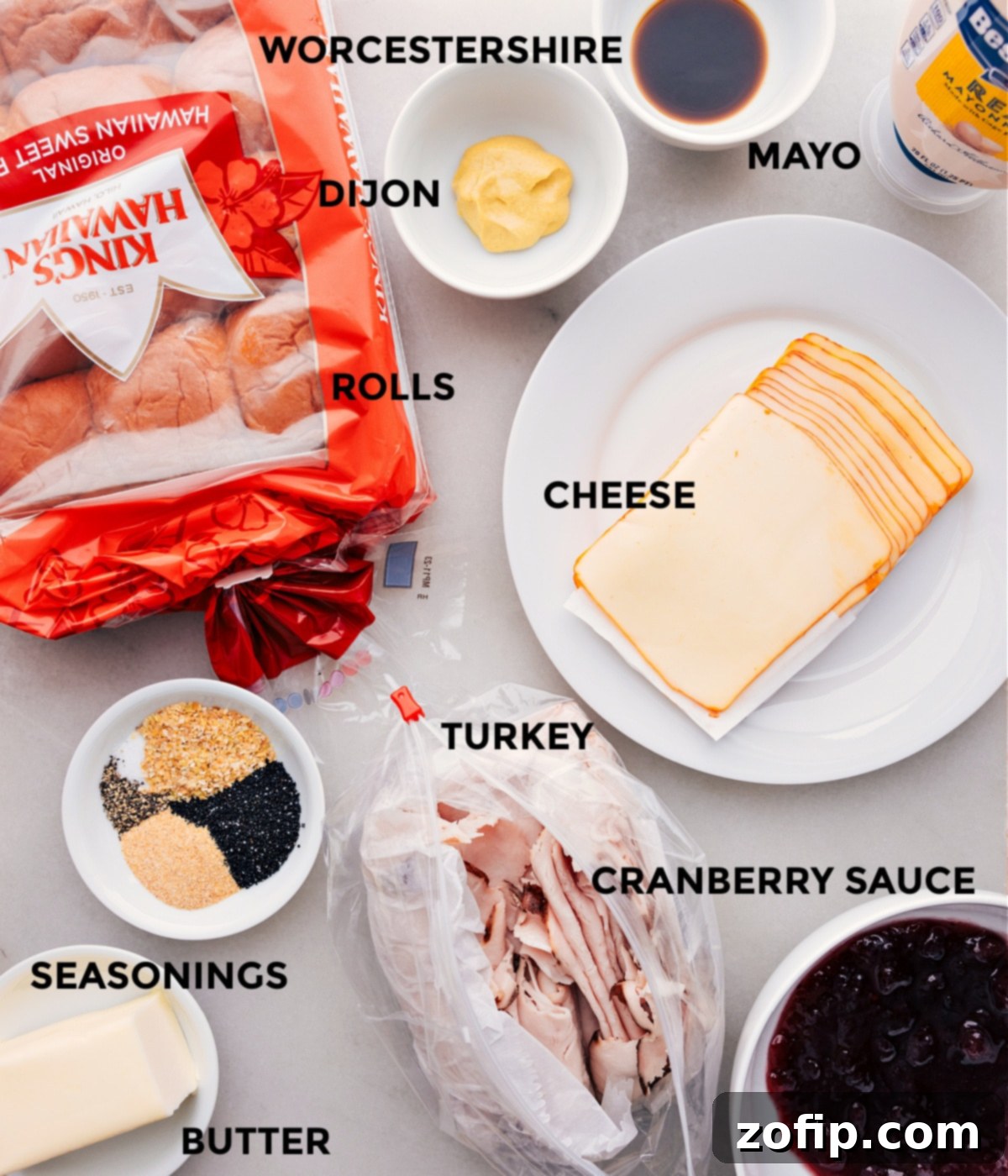 A vibrant display of all the essential ingredients for Turkey Cranberry Sliders, meticulously prepped for easy assembly. The setup includes fluffy Hawaiian rolls, thinly sliced deli turkey, creamy Muenster cheese, tangy cranberry sauce, smooth mayonnaise, and a separate bowl with the rich, seasoned butter topping mixture, alongside Dijon mustard and Worcestershire sauce.