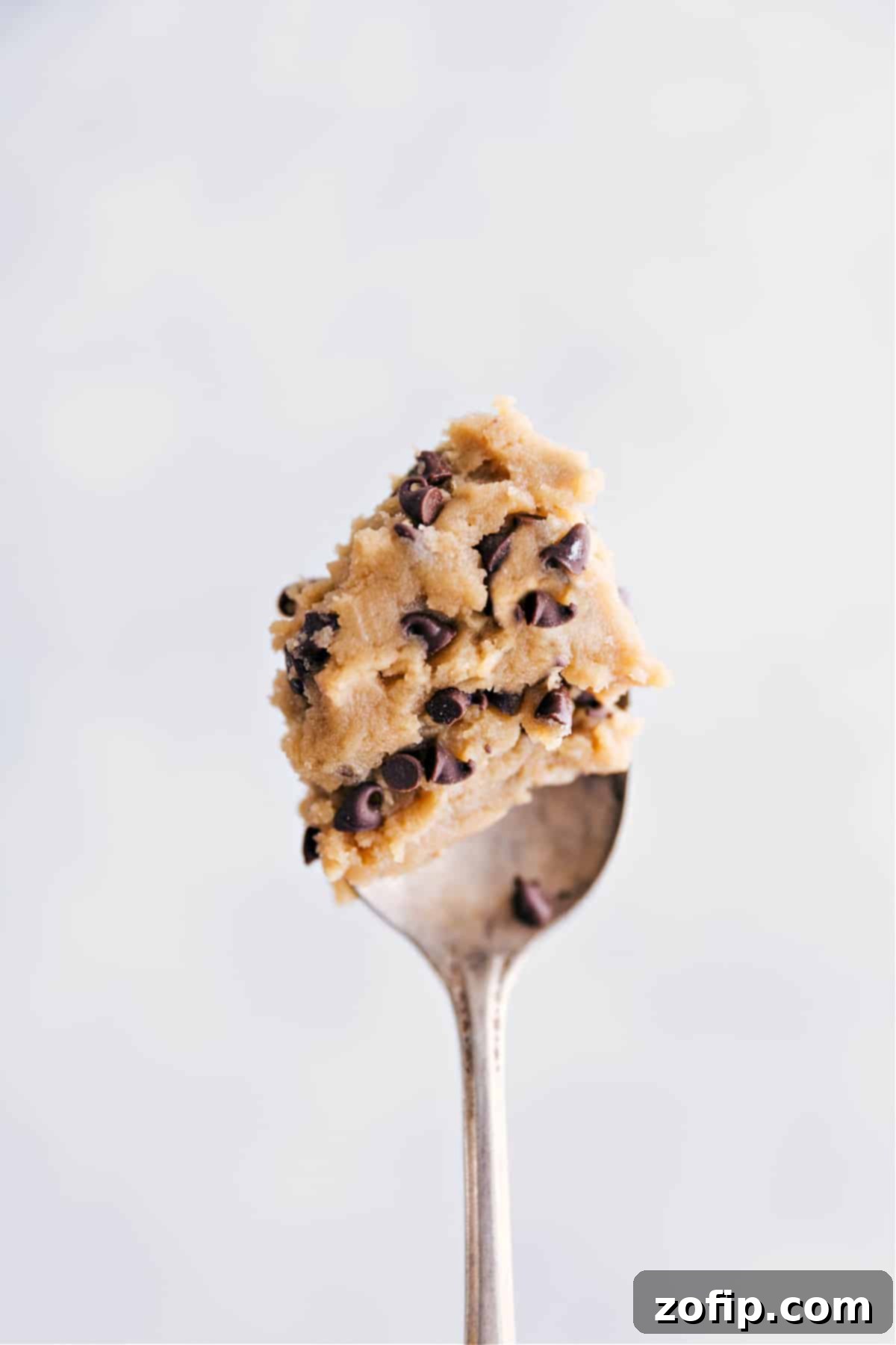 A single spoonful of perfectly textured edible chocolate chip cookie dough is held up, glistening with mini chocolate chips, against a softly blurred background, inviting immediate indulgence.
