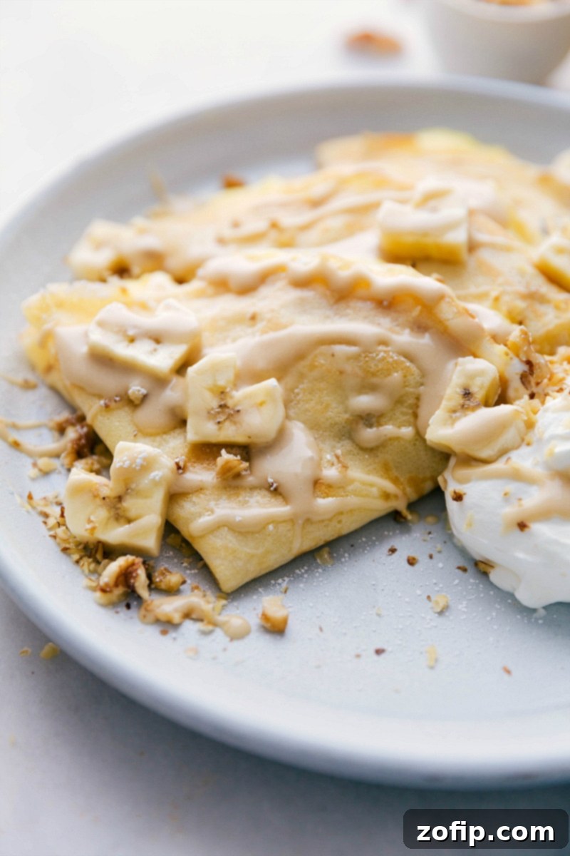 Overhead image of beautifully plated, ready-to-eat Banana Crepes, garnished with fresh banana slices, chopped walnuts, and a dusting of powdered sugar. The golden crepes are folded into triangles, inviting a delicious first bite.