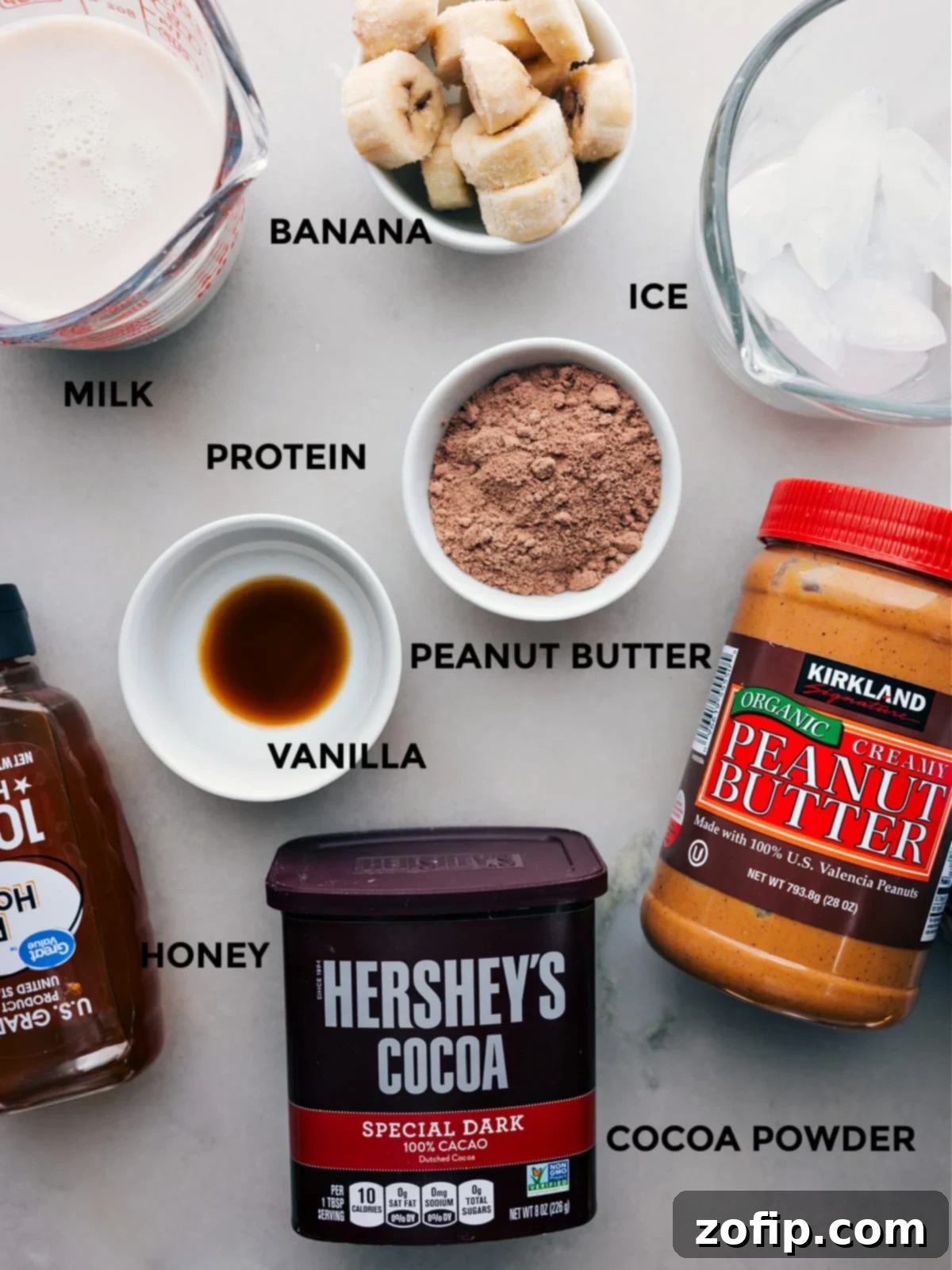 Ingredients In Peanut Butter Protein Shake Recipe All the ingredients including cocoa powder, vanilla, peanut butter, protein powder, bananas, milk, ice, and honey prepped out for easy assembly, displayed beautifully.
