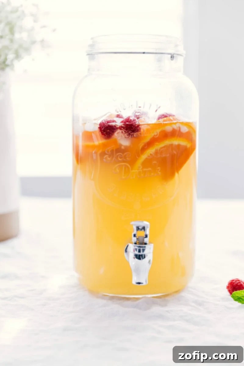 Ready-to-drink white grape punch in an elegant glass drink dispenser with fresh Cara Cara orange slices and raspberries inside, making it both delicious and visually beautiful for any party.