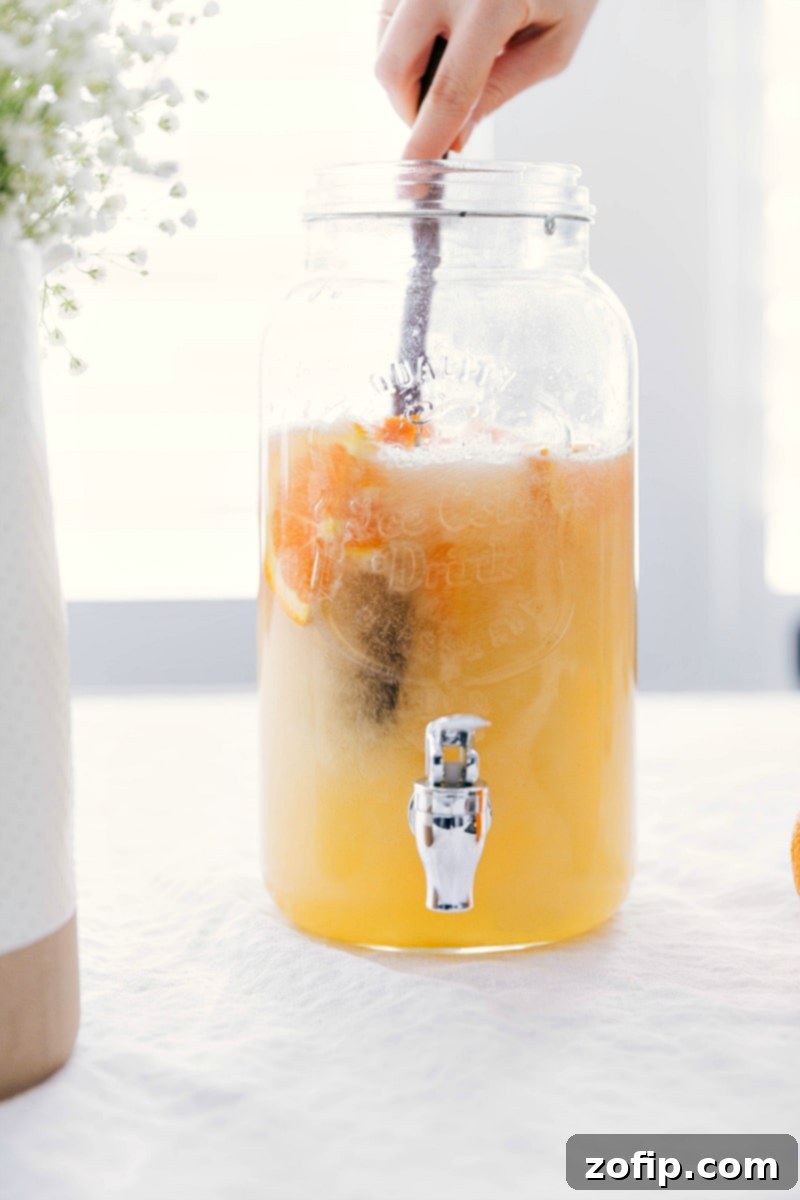 A refreshing and delicious white grape punch being gently stirred with a long spoon in a stylish glass serving canister, highlighting its inviting color and freshness.