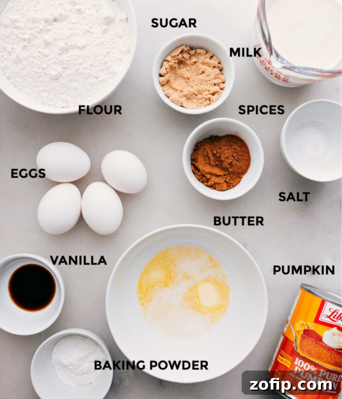 All the essential ingredients for pumpkin waffles laid out for preparation: flour, salt, baking powder, cinnamon, pumpkin pie spice, brown sugar, pumpkin puree, milk, vanilla extract, eggs, and butter.