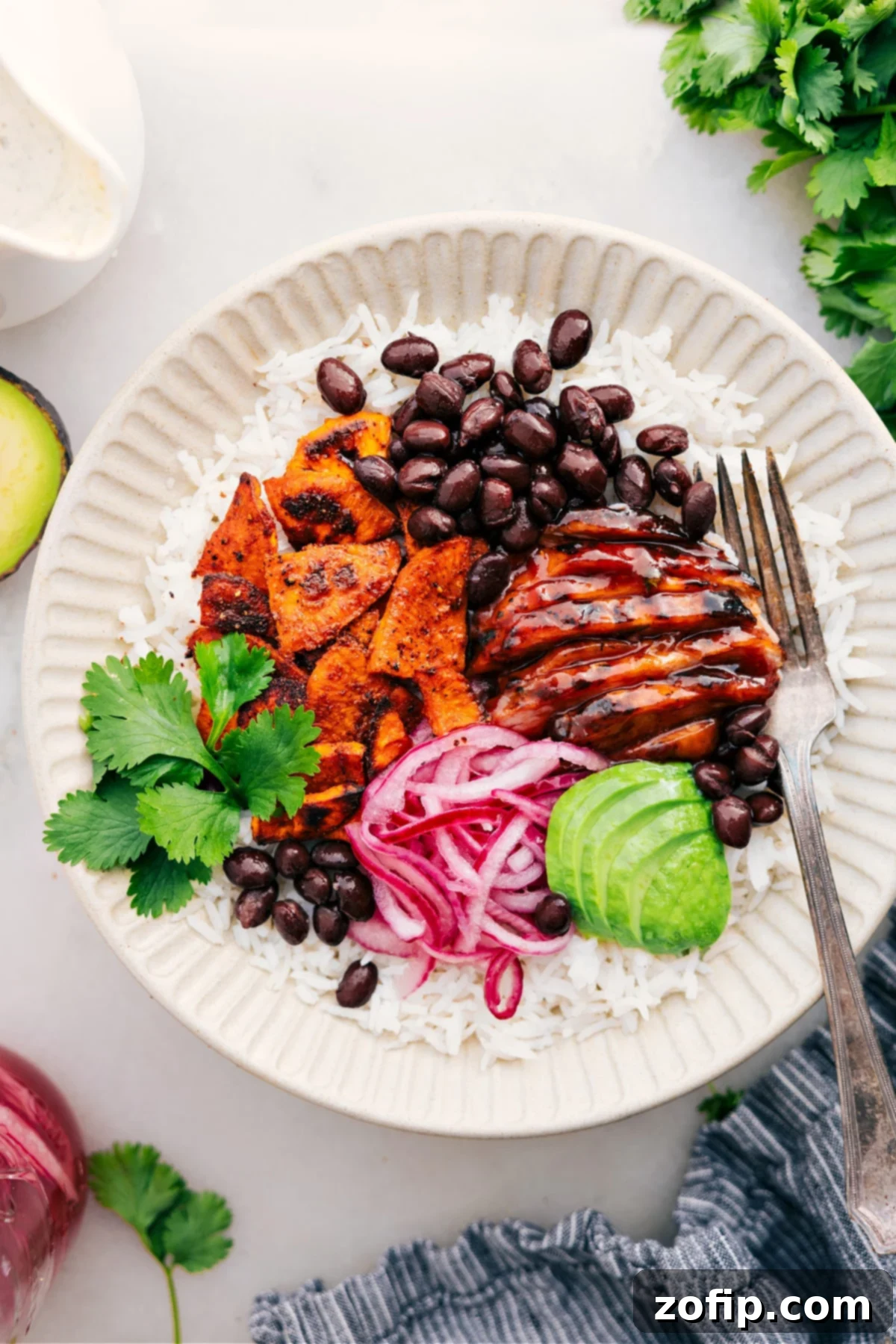 A colorful BBQ Chicken Bowl with rice, sweet potatoes, black beans, pickled onions, avocado, and BBQ chicken, drizzled with green goddess dressing.