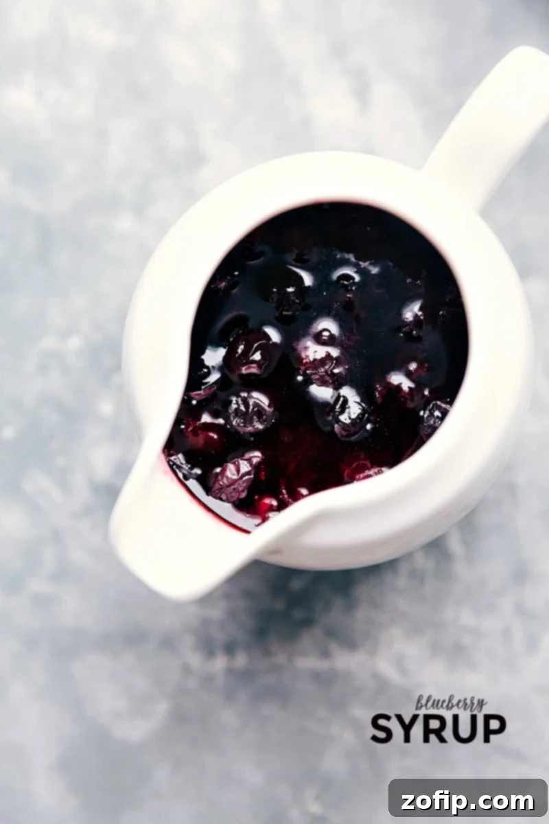 Fresh blueberries, a little bit of sugar, cornstarch, vanilla, and water make for the most incredible Blueberry Syrup! Ready-to-serve Blueberry Syrup in a glass pitcher, with fresh blueberries scattered around.