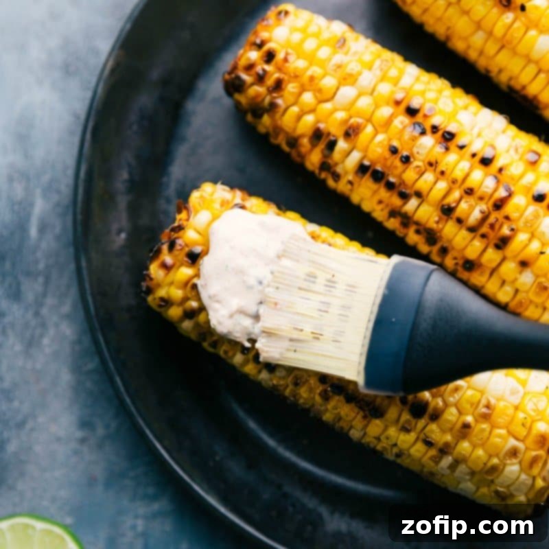 Applying creamy Elote sauce generously onto a perfectly roasted ear of corn using a pastry brush, highlighting the even coating and rich texture.