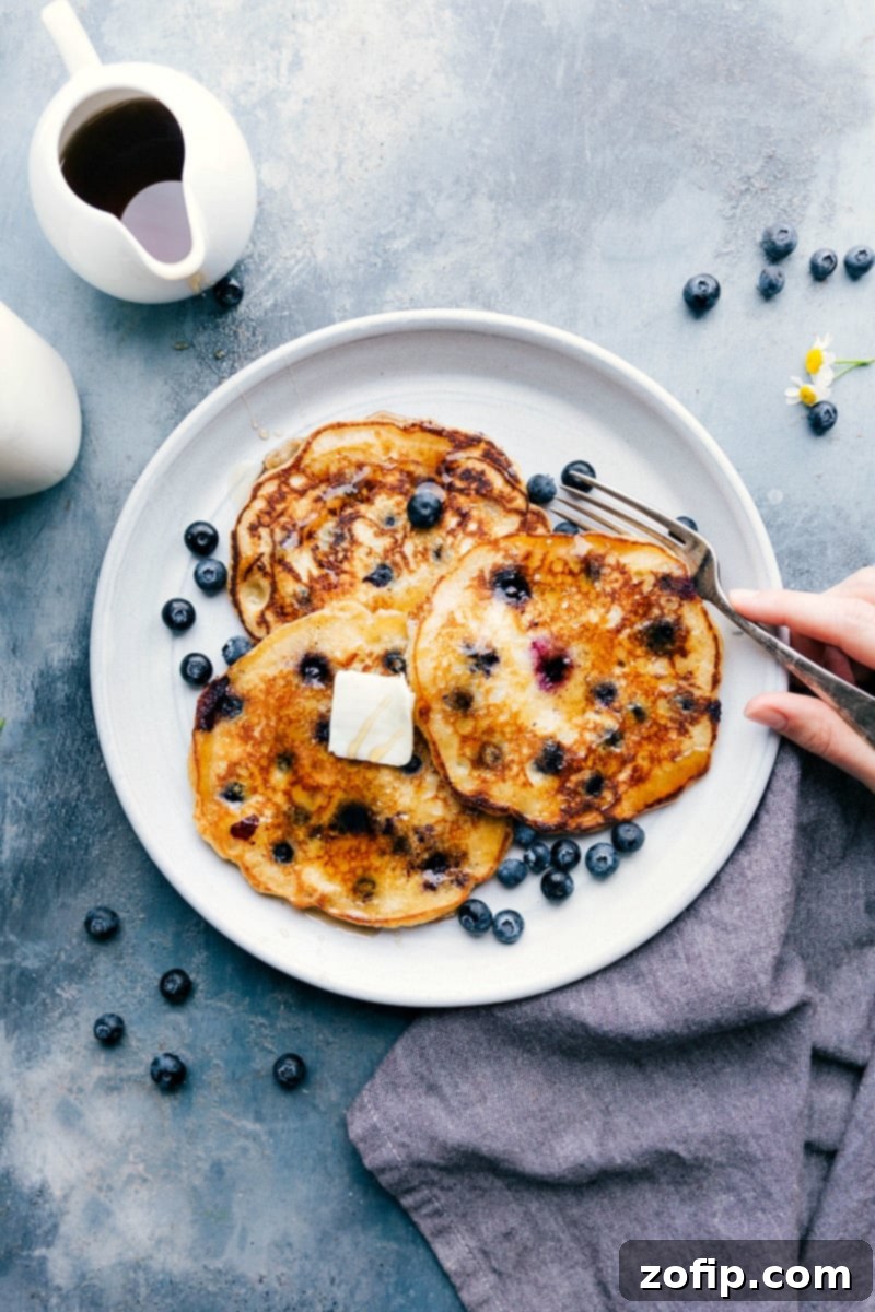 Blueberry Pancakes with Butter and Syrup A stack of warm blueberry pancakes on a plate, topped with melting butter and drizzled with syrup, with a fork on the side ready for enjoying.